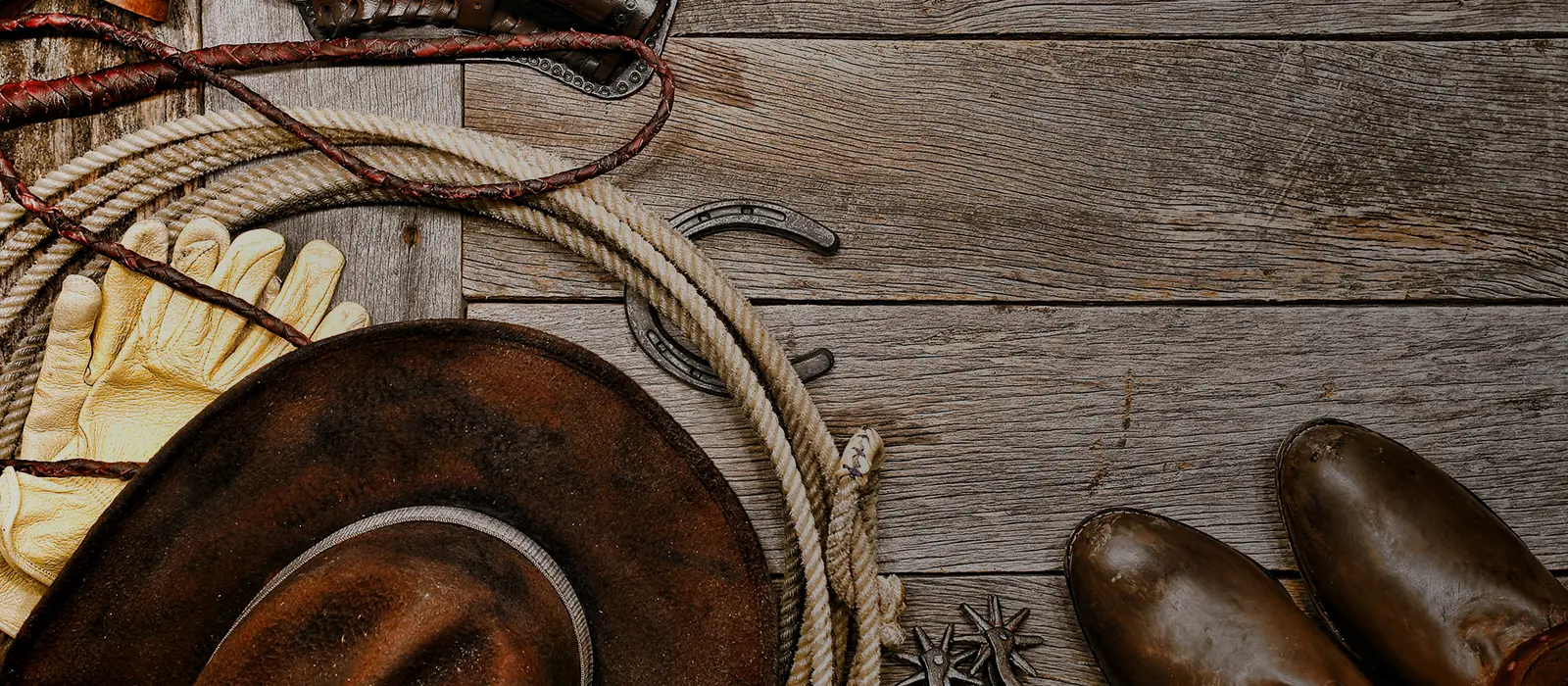 Cowboy hat, boots, working gloves, rope and horseshoe laying on weathered boards.